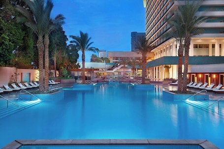 A hotel pool area with a clear view of the pool, lounge chairs, palm trees, and a building in the background.