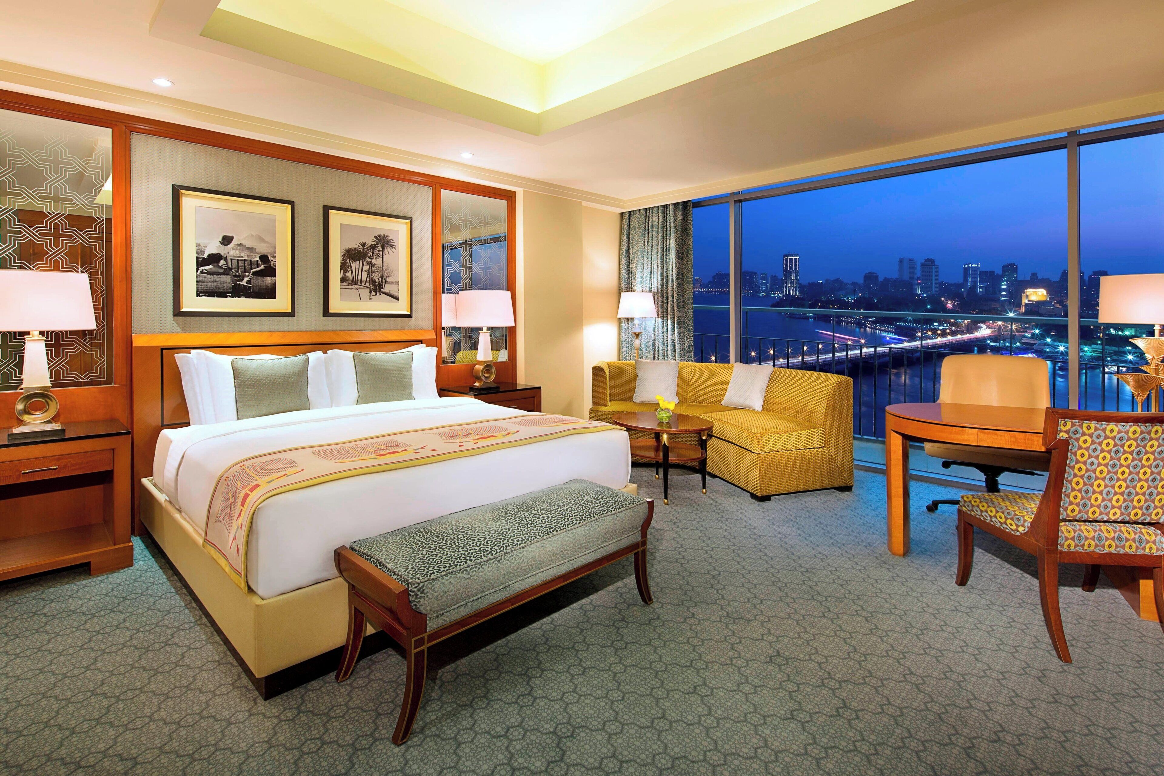 Habitación de hotel con una cama grande, un sofá, un escritorio y vista a la ciudad de noche.