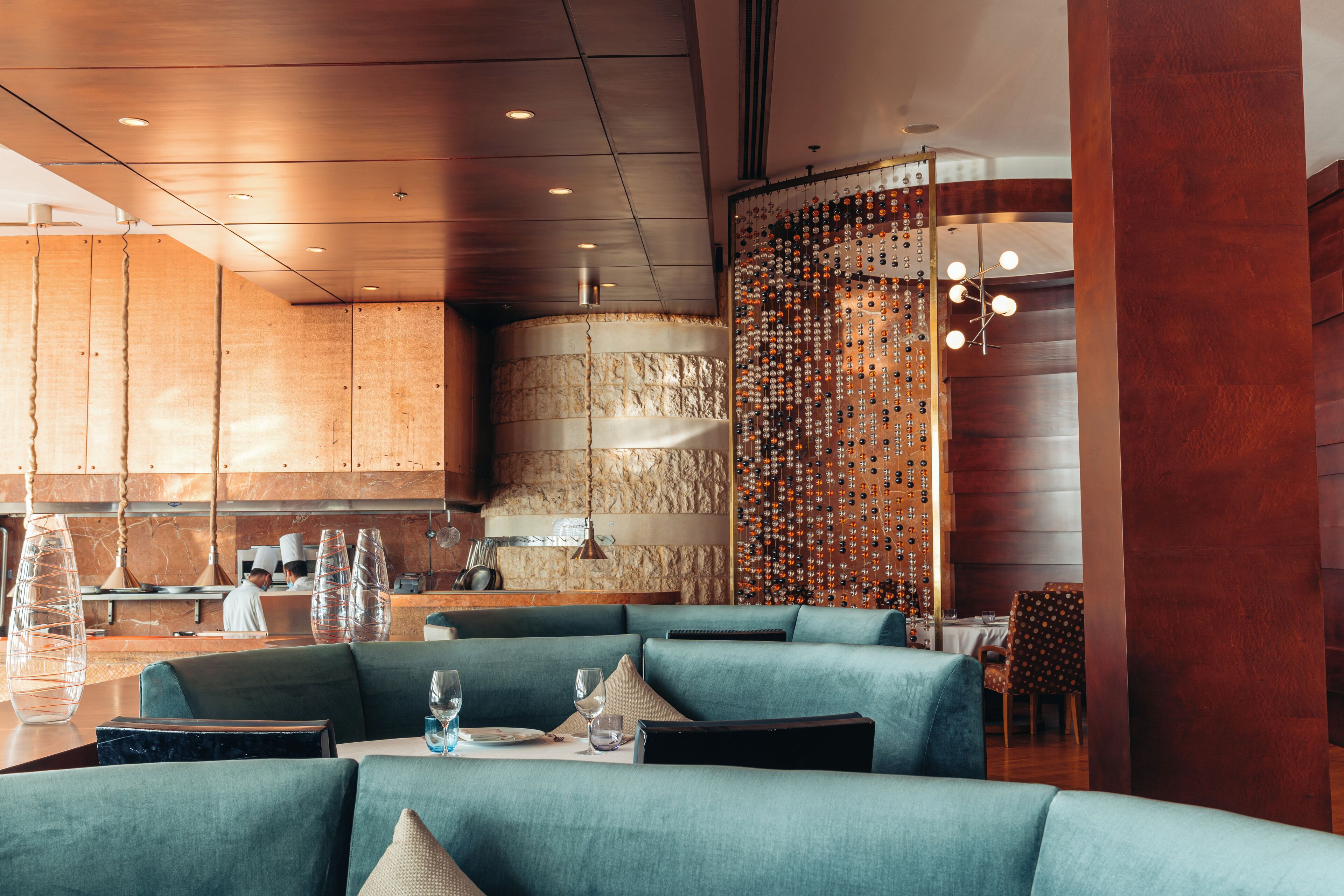 Un interior de restaurante moderno con cabinas tapizadas en color verde azulado, una pared de piedra y una estantería decorativa para vinos.