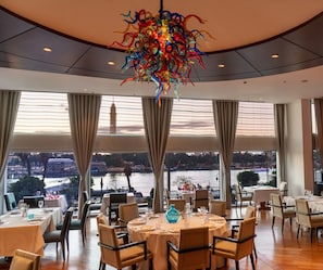 Un espace repas avec des tables dressées pour le repas, de grandes fenêtres offrant une vue sur une rivière et des bâtiments, et un lustre coloré.