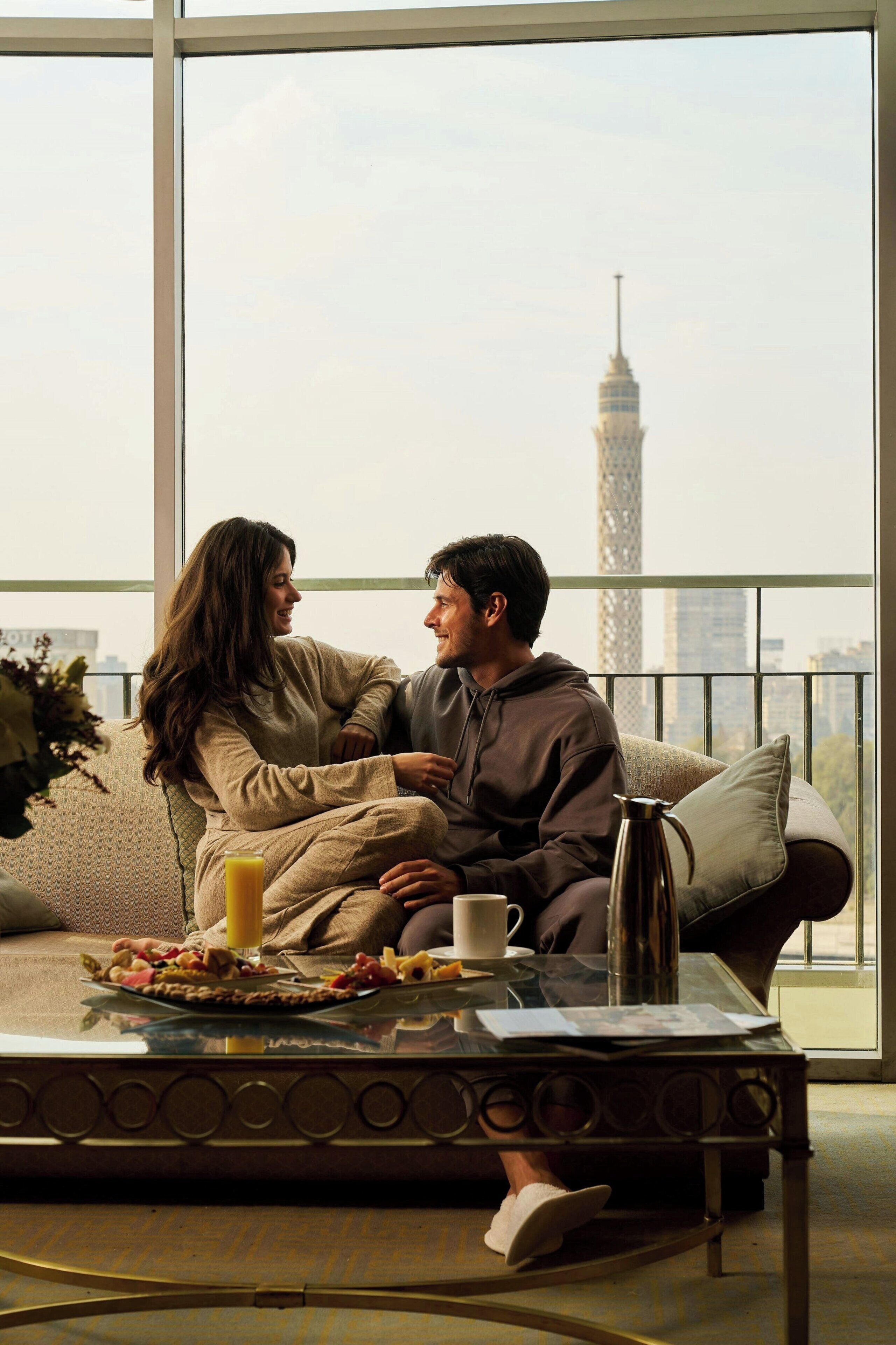 A couple sitting on a sofa with a cityscape view, a coffee table with breakfast items, and a large window.