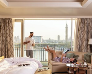 Una camera d'albergo con un letto, un divano, un balcone vista città e un tavolo da pranzo con prodotti per la colazione.