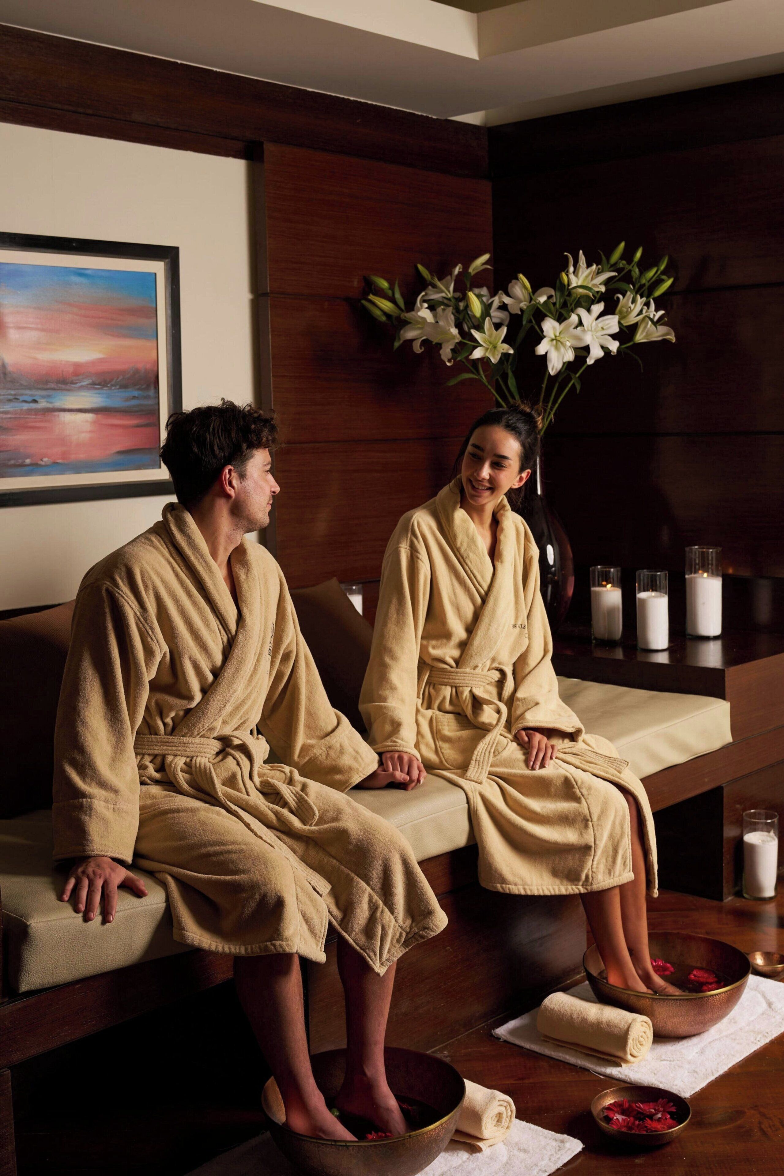 Ein Spa-Ambiente mit zwei Personen in Bademänteln, einer hölzernen Bank, einer Blumenschale, Kerzen und einem Gemälde an der Wand.