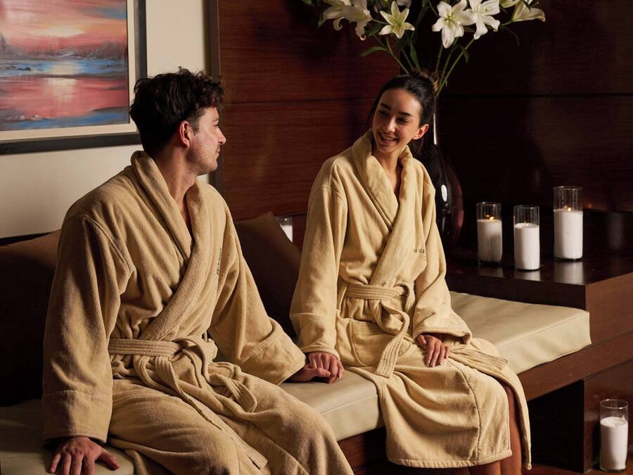 Un ambiente de spa con dos personas en batas, un banco de madera, un cuenco de flores, velas y un cuadro en la pared.
