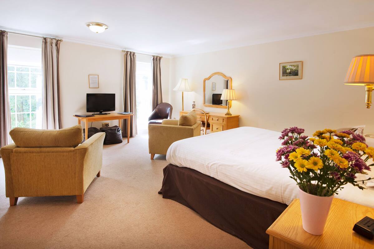 A hotel room with a bed, two armchairs, a desk with a television, a dresser, a mirror, and a vase of flowers.