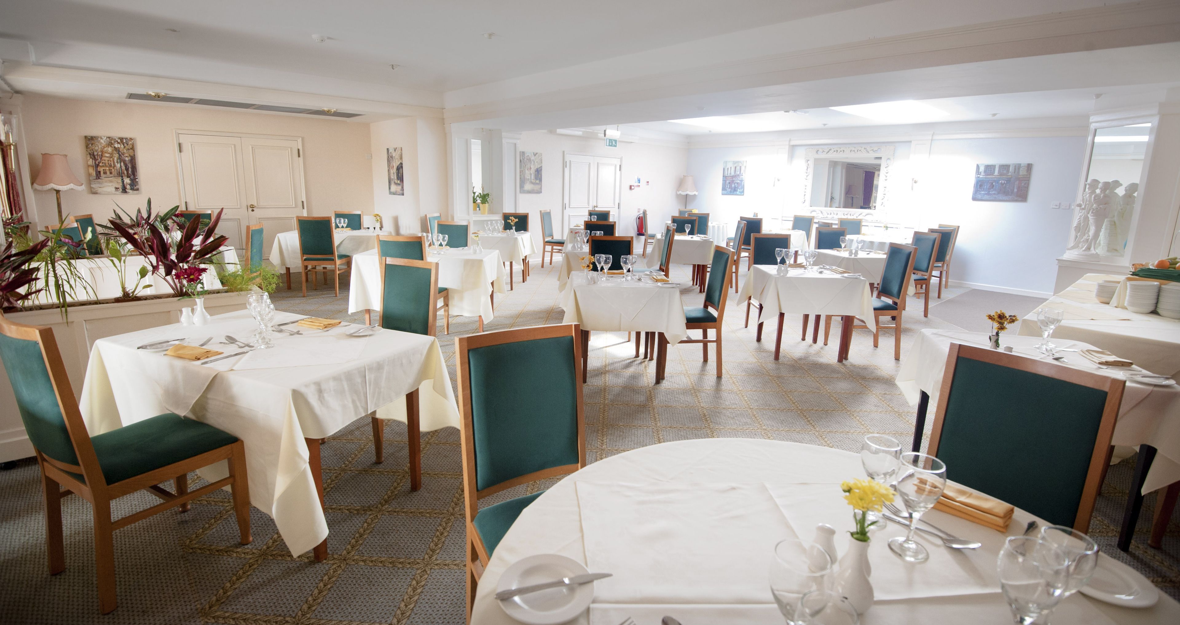 A spacious dining area with multiple tables and chairs, well-lit with natural light, and decorated with plants and artwork.