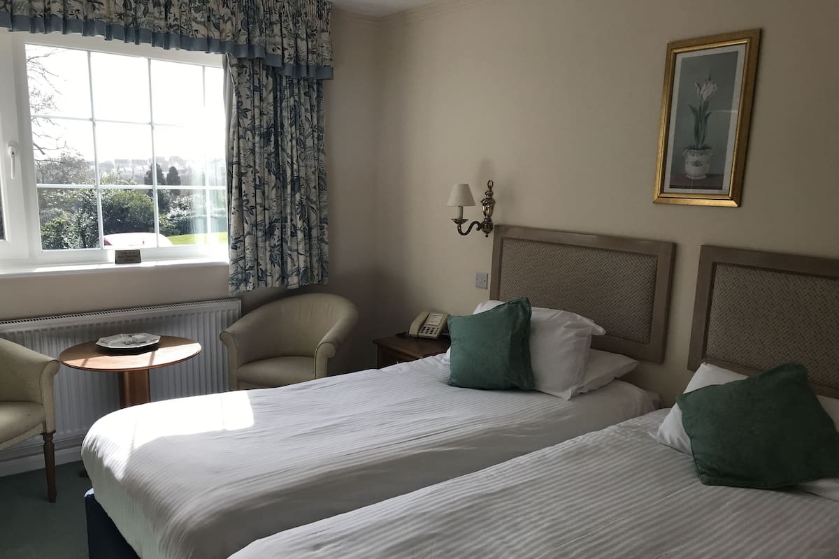 A hotel room with two beds, a chair, a small table, a lamp, and a window with curtains.