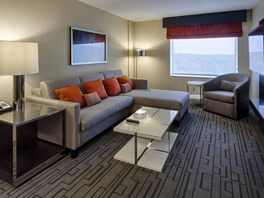 A modern hotel room with a sofa, armchair, glass coffee table, and a small side table with a lamp.