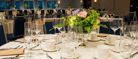 Un comedor con mesas redondas dispuestas para una comida formal, con manteles blancos, platos, cubiertos y copas de vino.