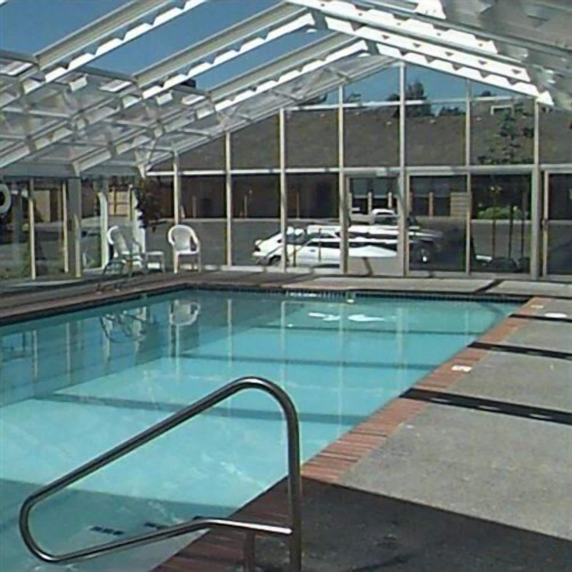 A swimming pool with a glass enclosure, a metal pool ladder, and a view of parked cars and a building in the background.