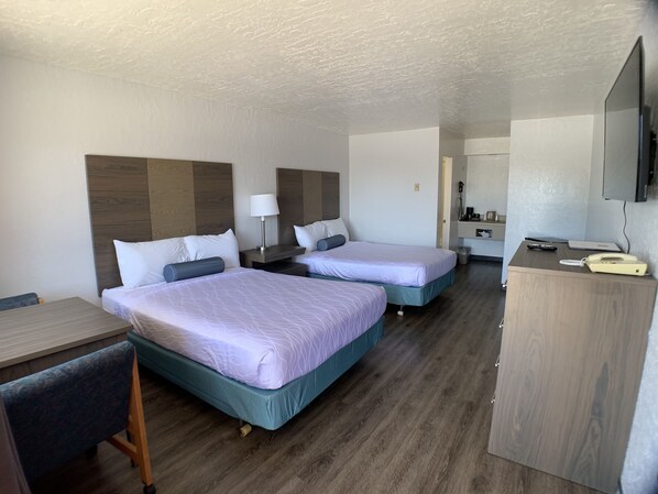 A hotel room with two beds, a desk, a chair, a TV, and a small kitchenette.