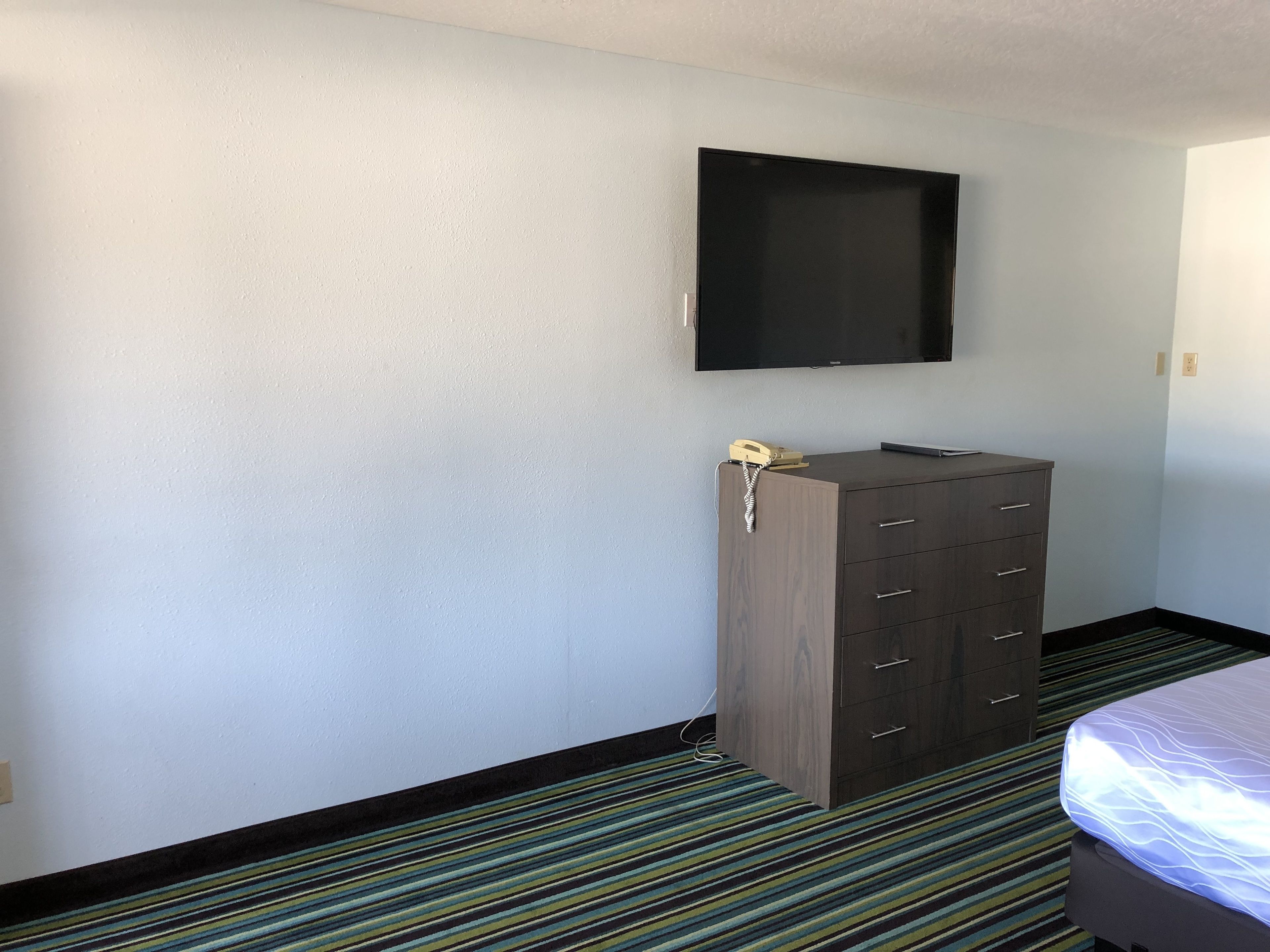A hotel room with a bed, a dresser, a flat-screen TV mounted on the wall, and a striped carpet.