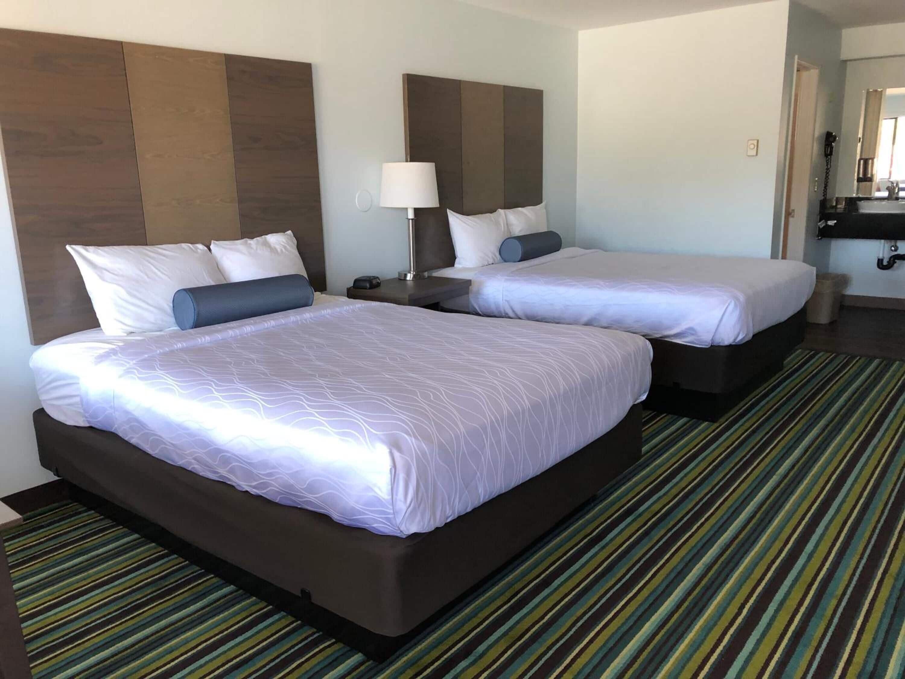 A hotel room with two beds, a nightstand, a lamp, and a bathroom visible through an open door.