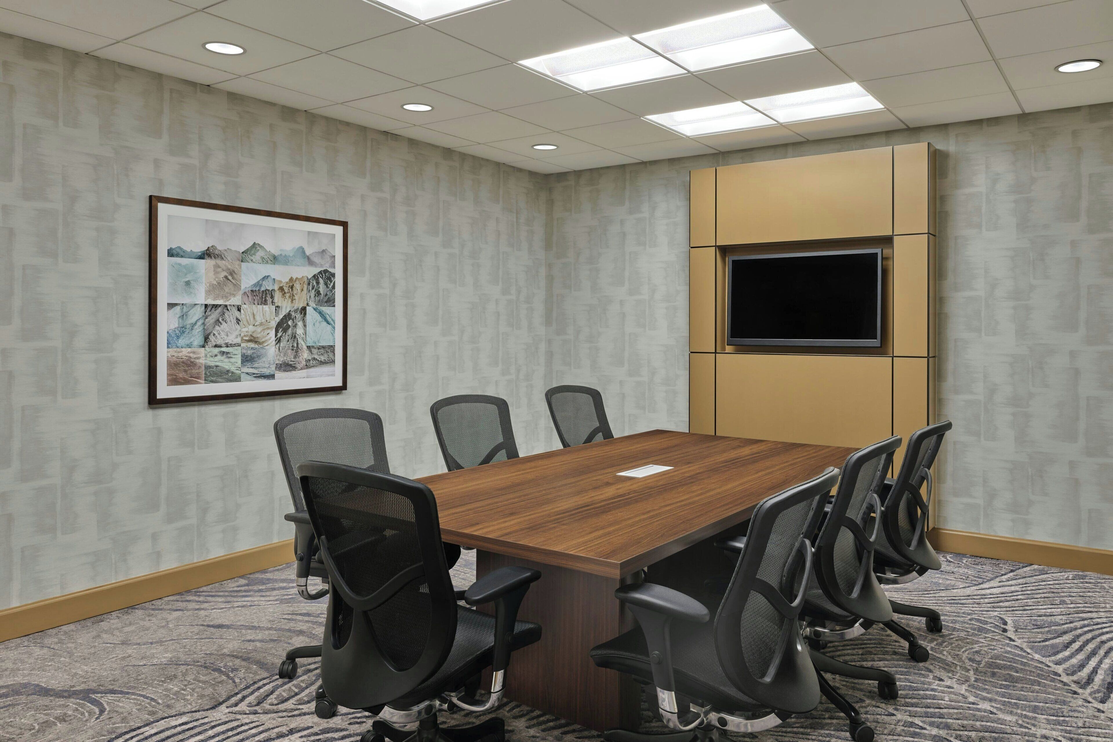 A conference room with a wooden table, six office chairs, a mounted TV, and a framed picture on the wall.