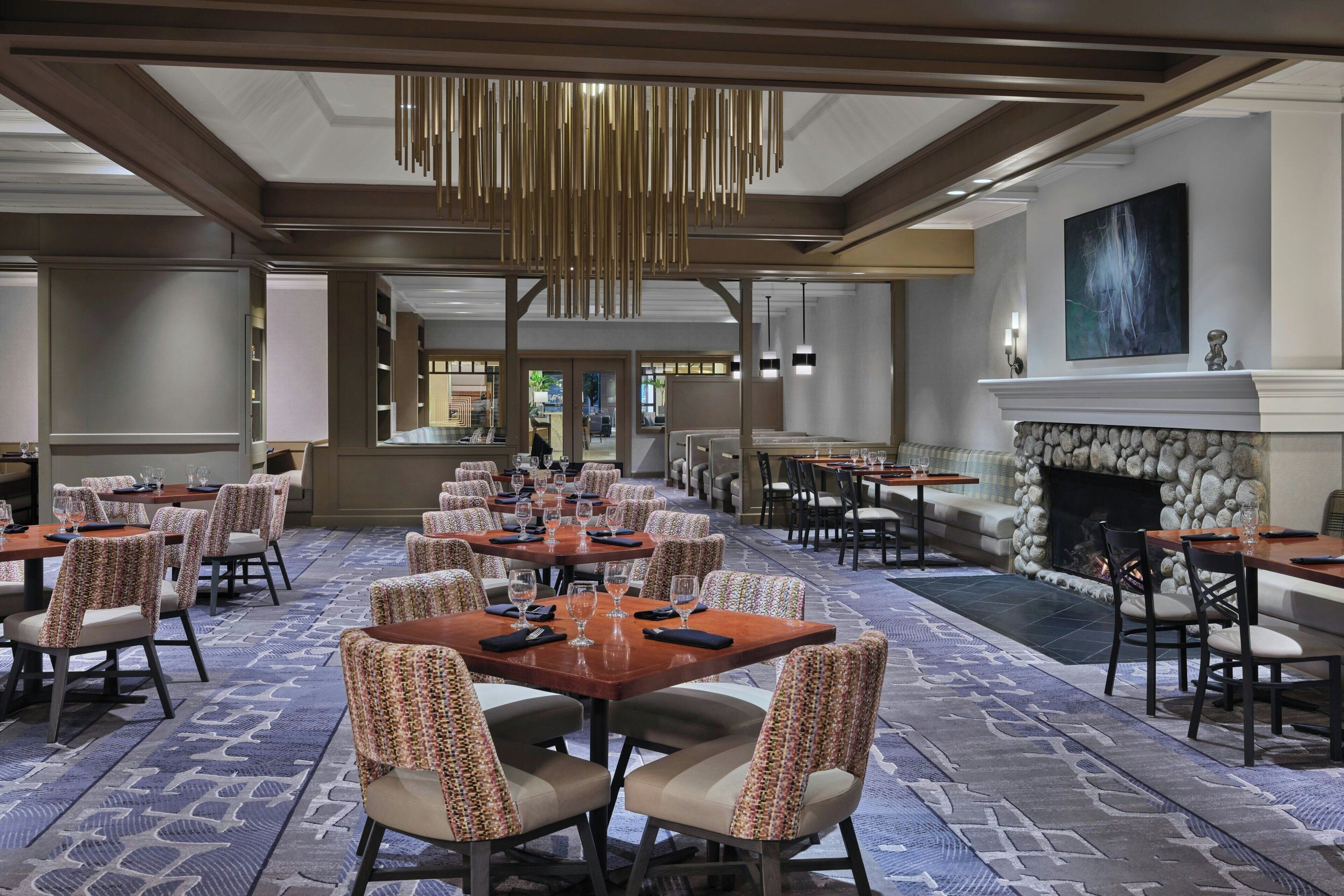 A spacious dining area with multiple tables and chairs, a stone fireplace, and a modern chandelier.