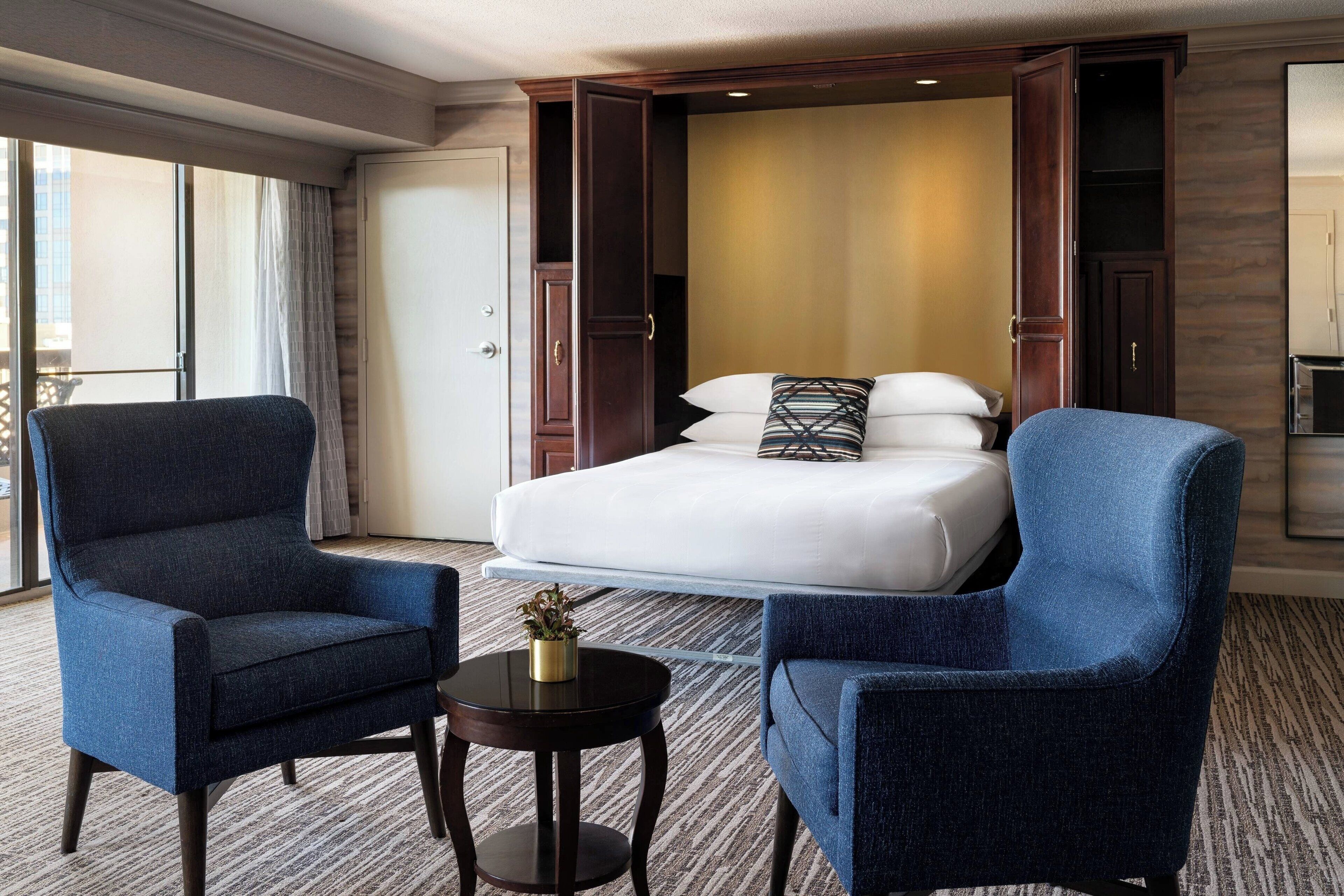 A hotel room with a large bed, two armchairs, a small table, and a view of the city through large windows.