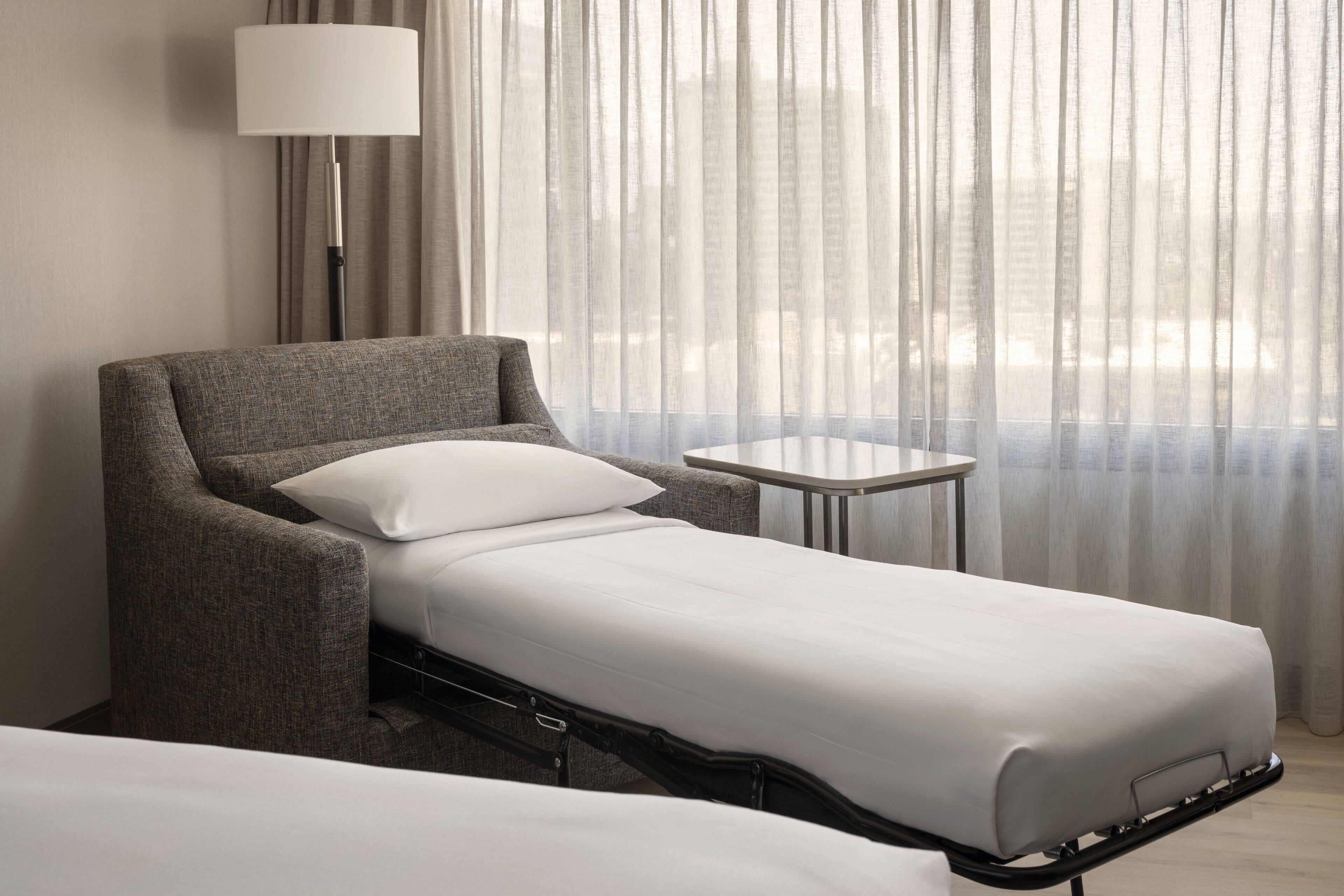 Habitación de hotel con una cama individual, una mesita pequeña, una lámpara y vistas a través de cortinas translúcidas.