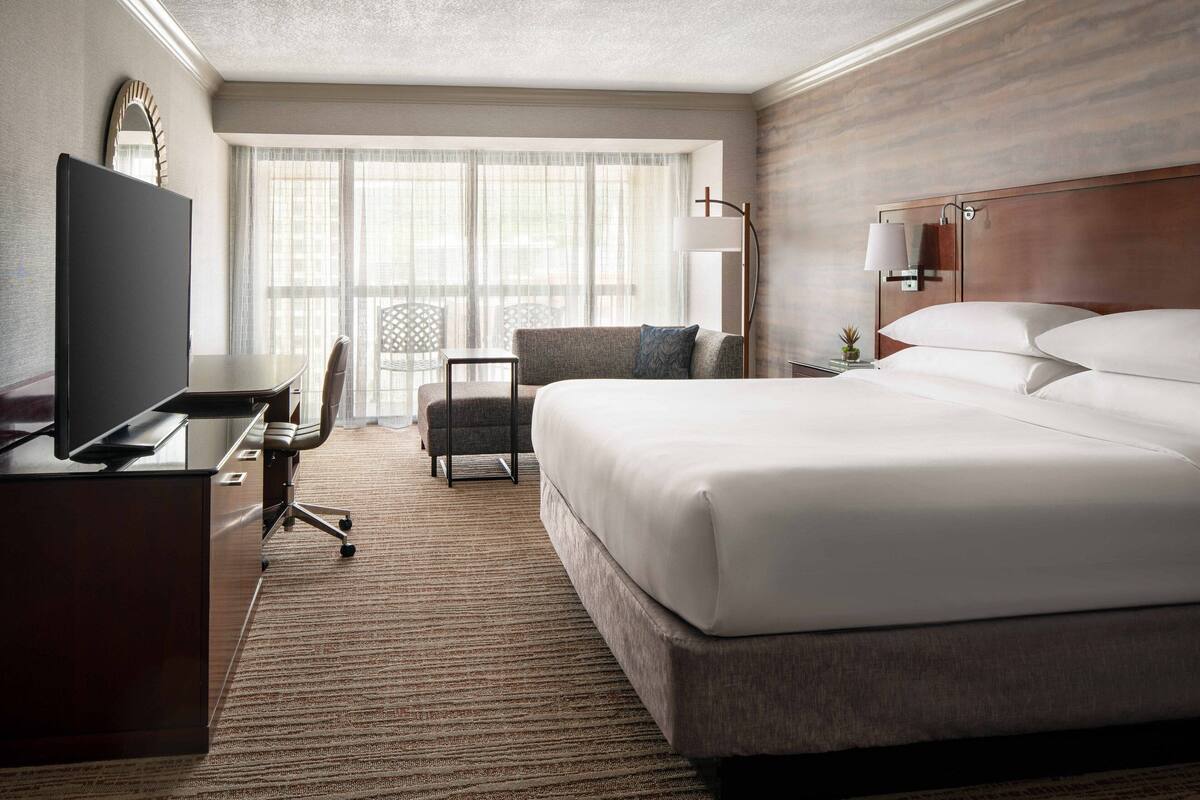 Quarto de hotel com uma cama grande, uma escrivaninha com televisão de tela plana, uma cadeira, uma mesinha e uma janela ampla com cortinas translúcidas.