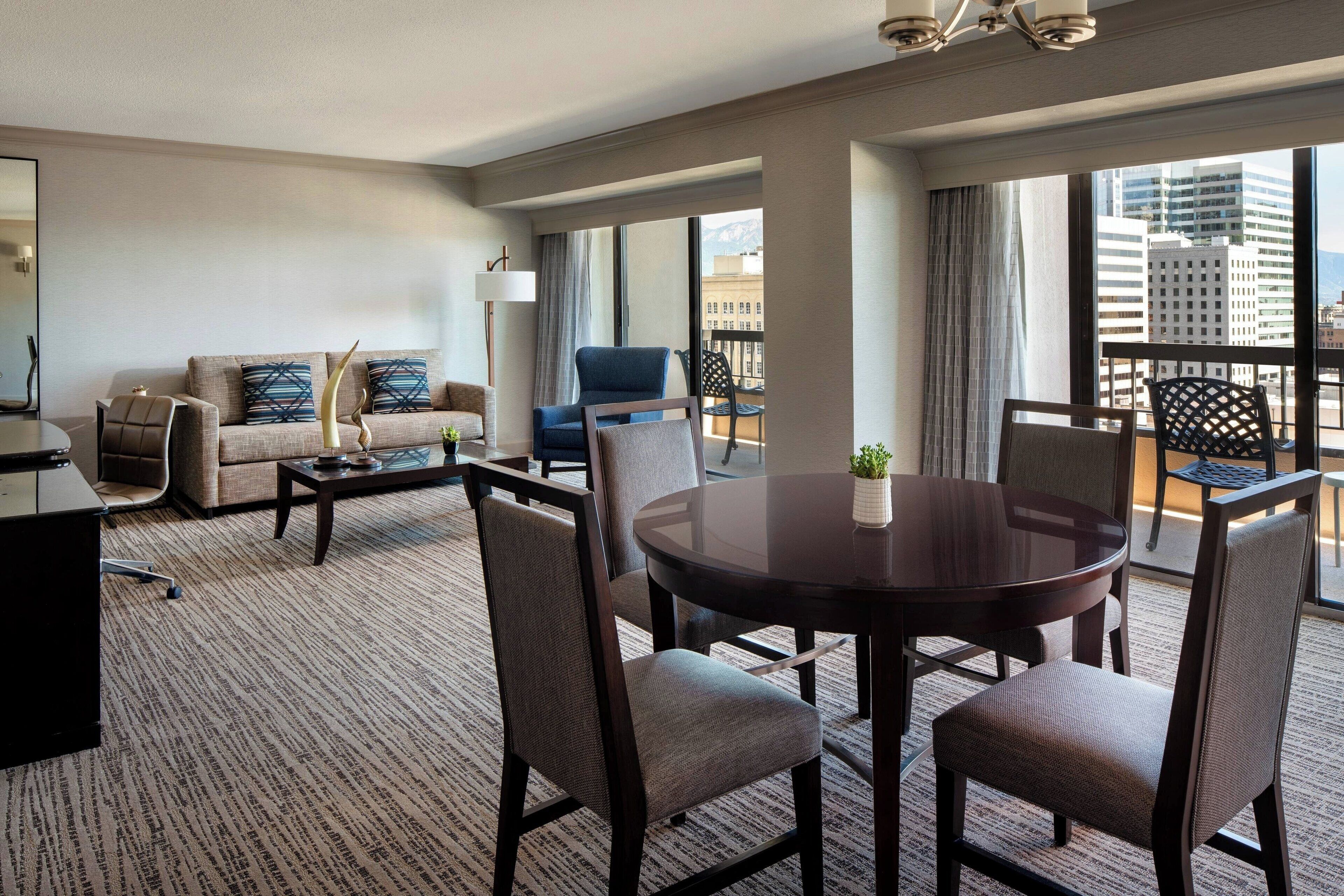 A hotel room with a sofa, armchairs, a dining table, and a balcony with a view of the city.