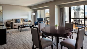 A hotel room with a sofa, armchairs, a dining table, and a balcony with a view of the city.