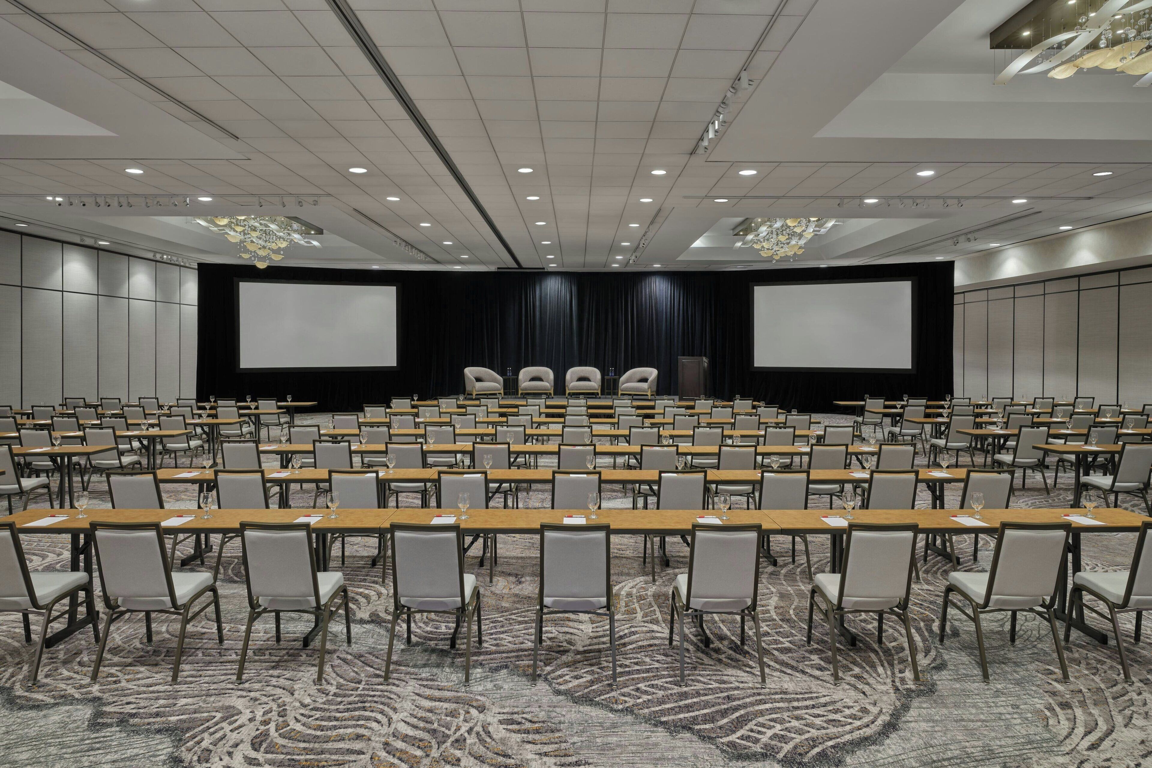 Un salón de conferencias con filas de mesas y sillas, una pantalla grande y un escenario con cortinas.