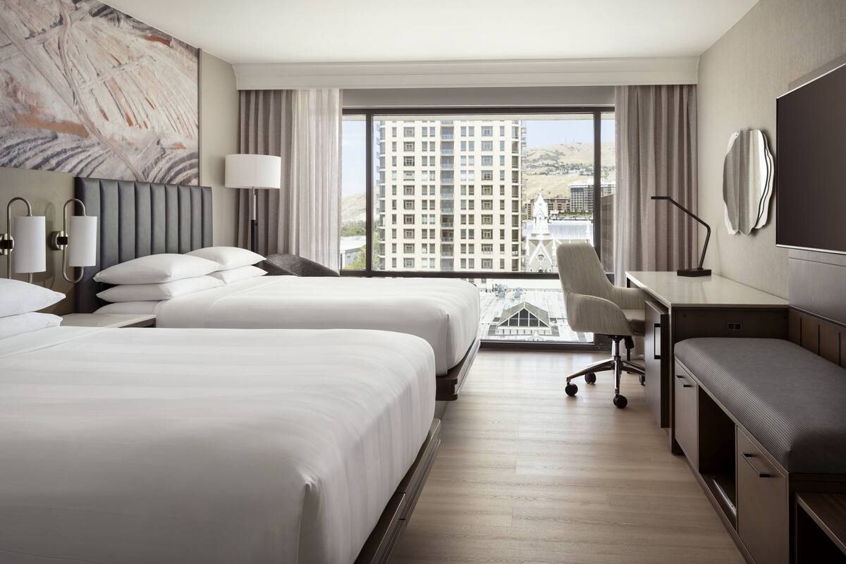 Quarto de hotel com duas camas, uma mesa de trabalho, uma cadeira, televisão e uma janela grande com vista para a cidade.
