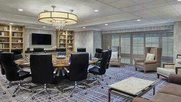 A conference room with a round table, black chairs, a chandelier, a bookshelf, and a large window with blinds.
