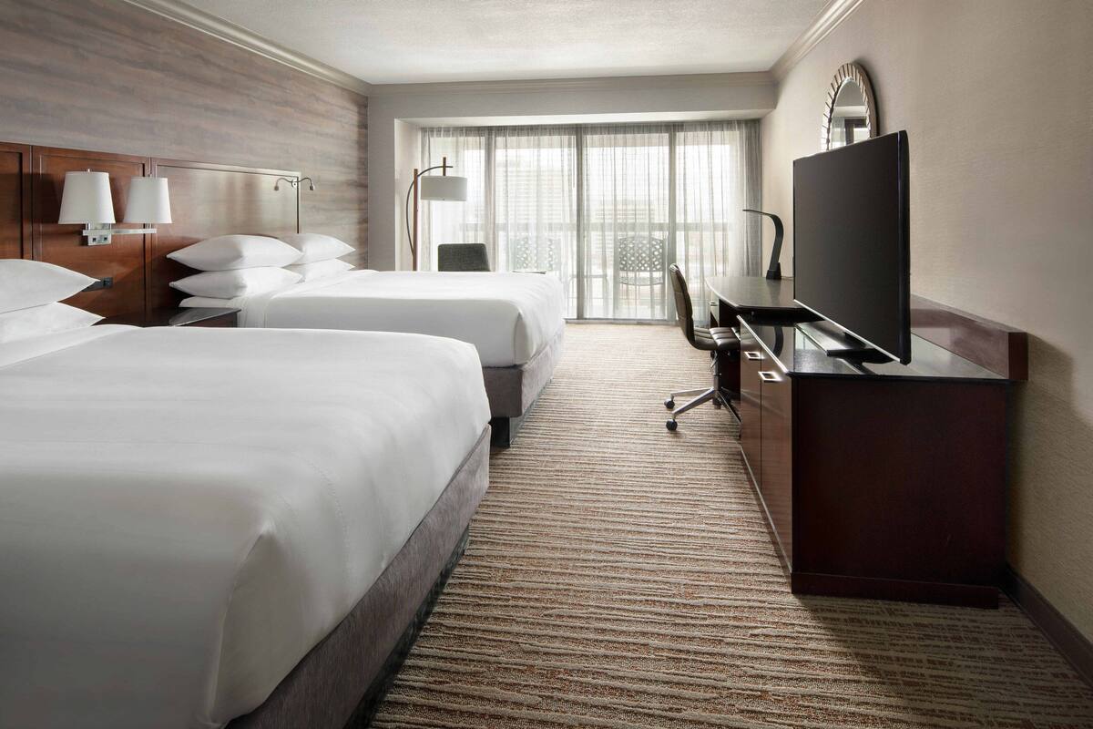 Quarto de hotel com duas camas, uma escrivaninha com televisão de tela plana, uma cadeira e uma janela grande com cortinas leves.