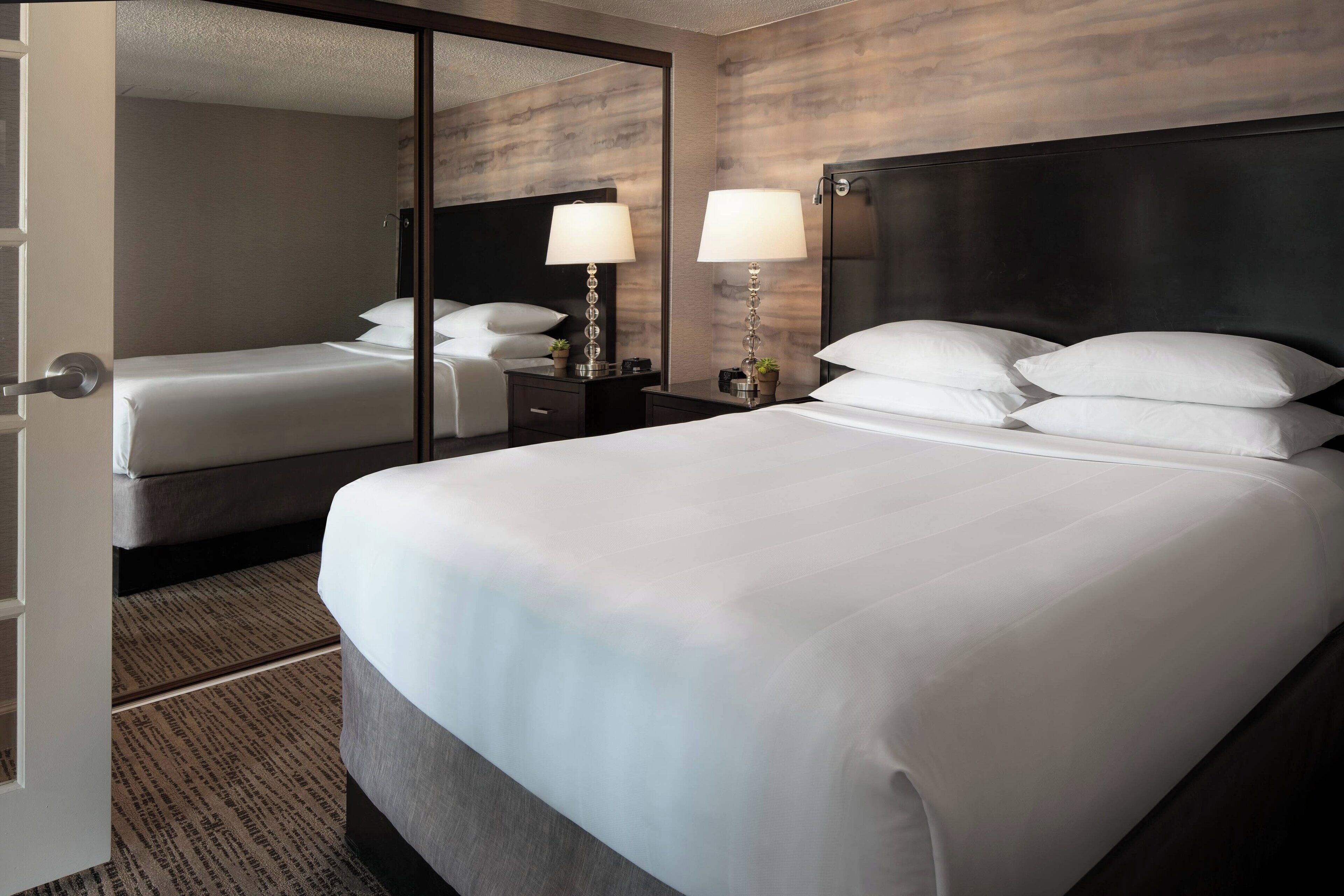 A hotel room with a large bed, a nightstand with two lamps, a mirror, and a wardrobe.