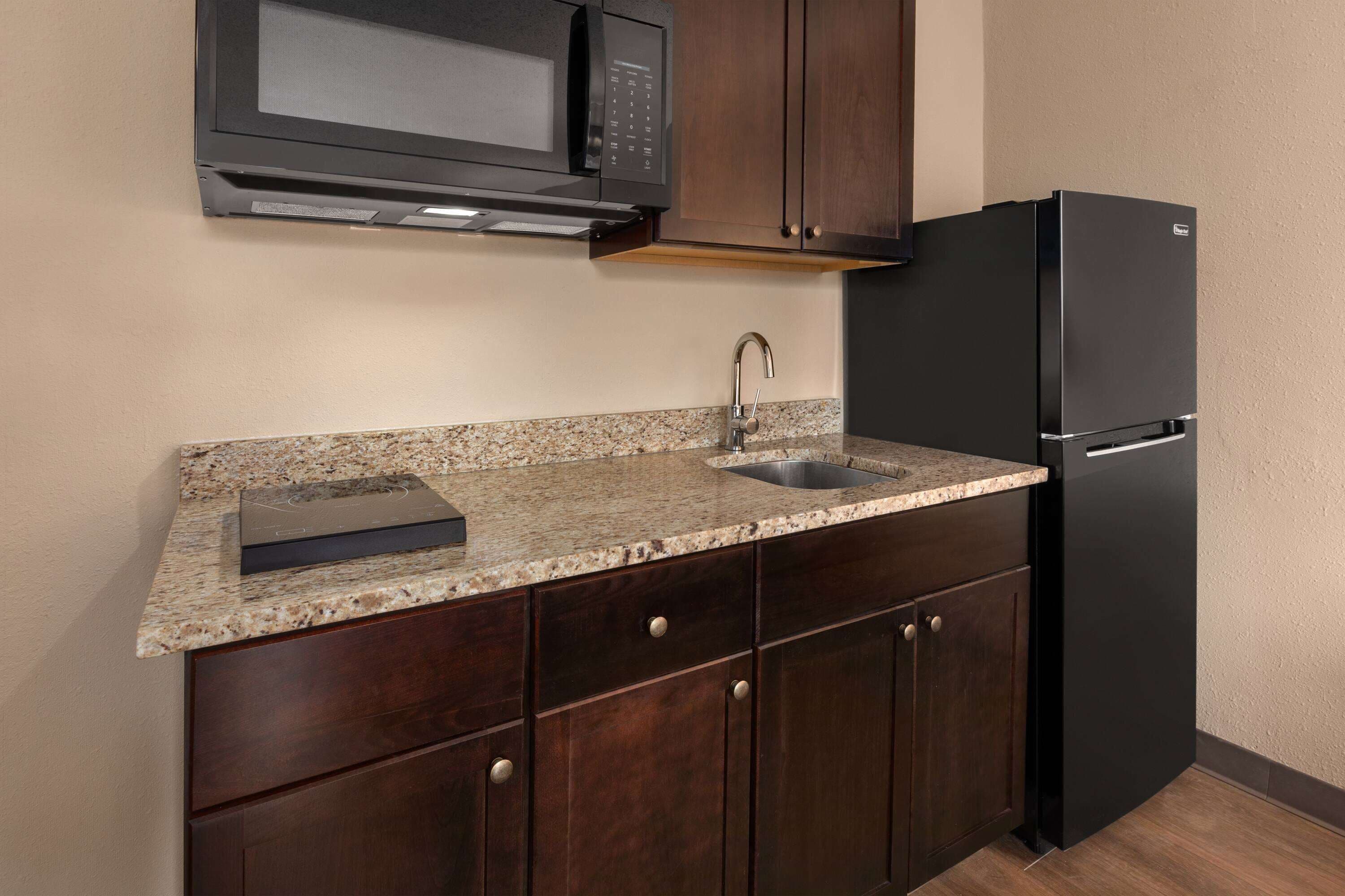 Cocina con armarios de madera oscura, encimera de granito, pileta de acero inoxidable, refrigerador negro y microondas sobre la encimera.