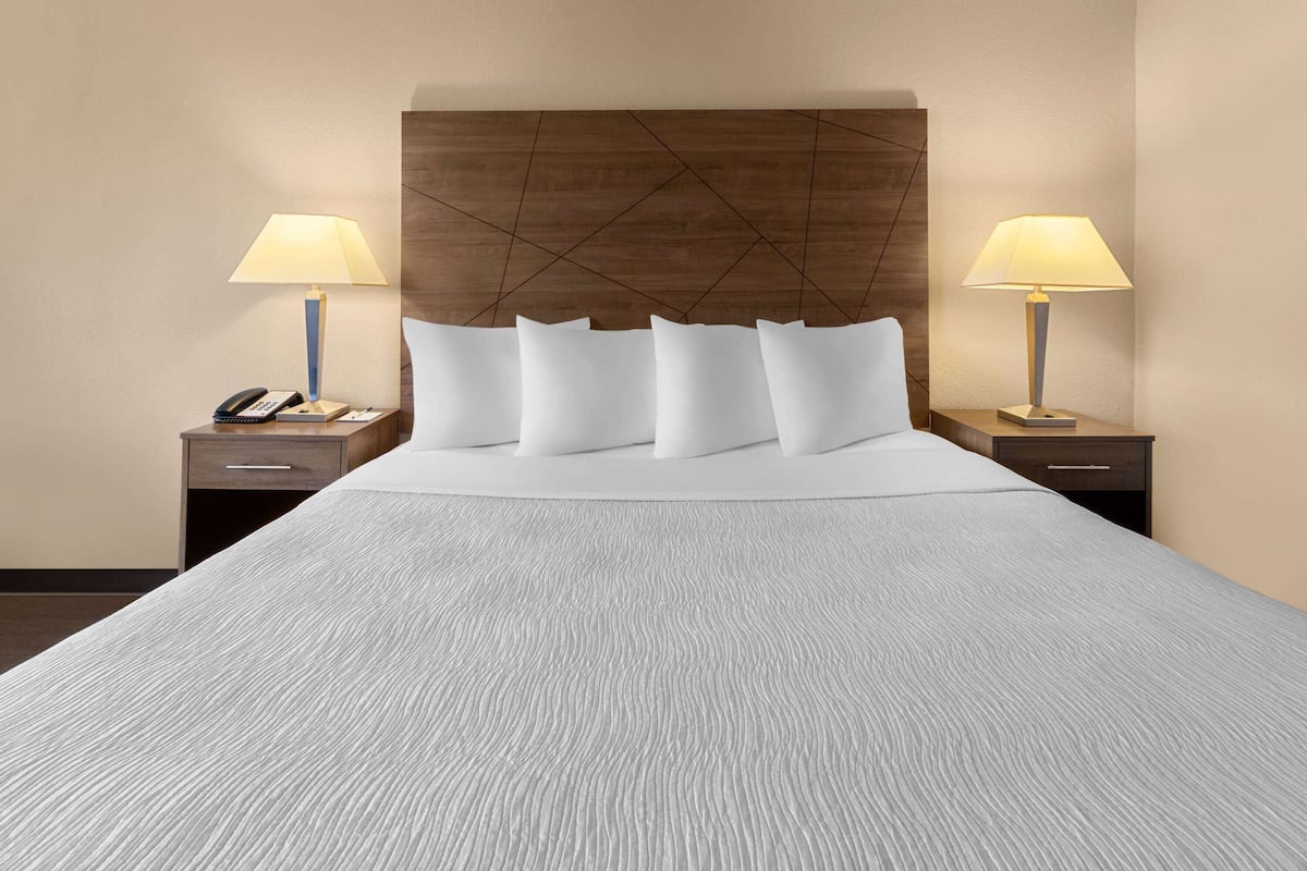 Una cama bien hecha con una colcha blanca texturizada, un cabecero de madera con diseño geométrico, dos mesitas de noche con lámparas, y un teléfono en una de las mesitas.