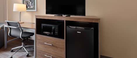 A hotel room with a flat-screen TV mounted above a wooden cabinet, a microwave, and a refrigerator. There is a desk with a chair and a lamp.