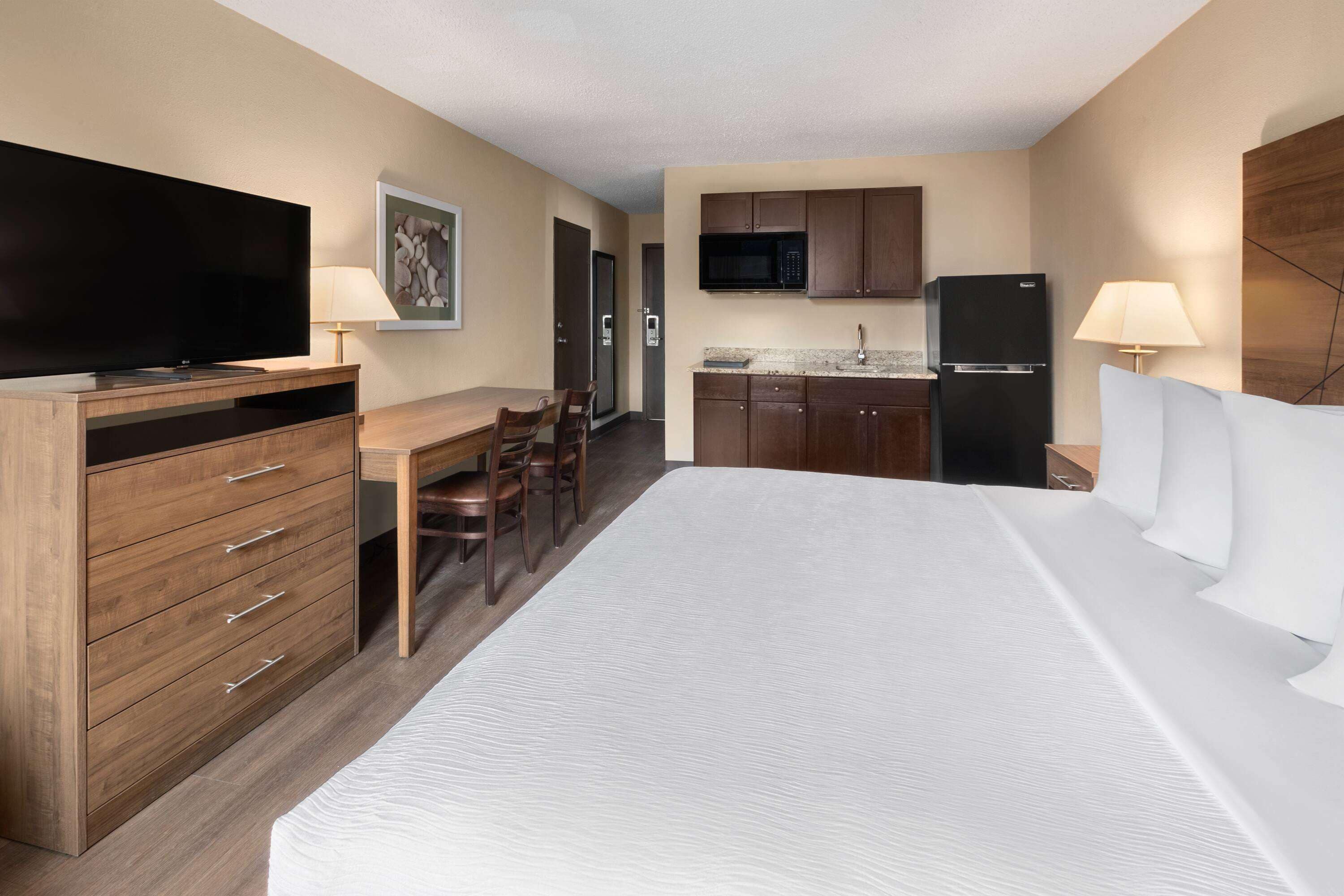 Habitación de hotel con cama, televisor, escritorio, sillas y zona de cocina con microondas, refrigerador y armarios.