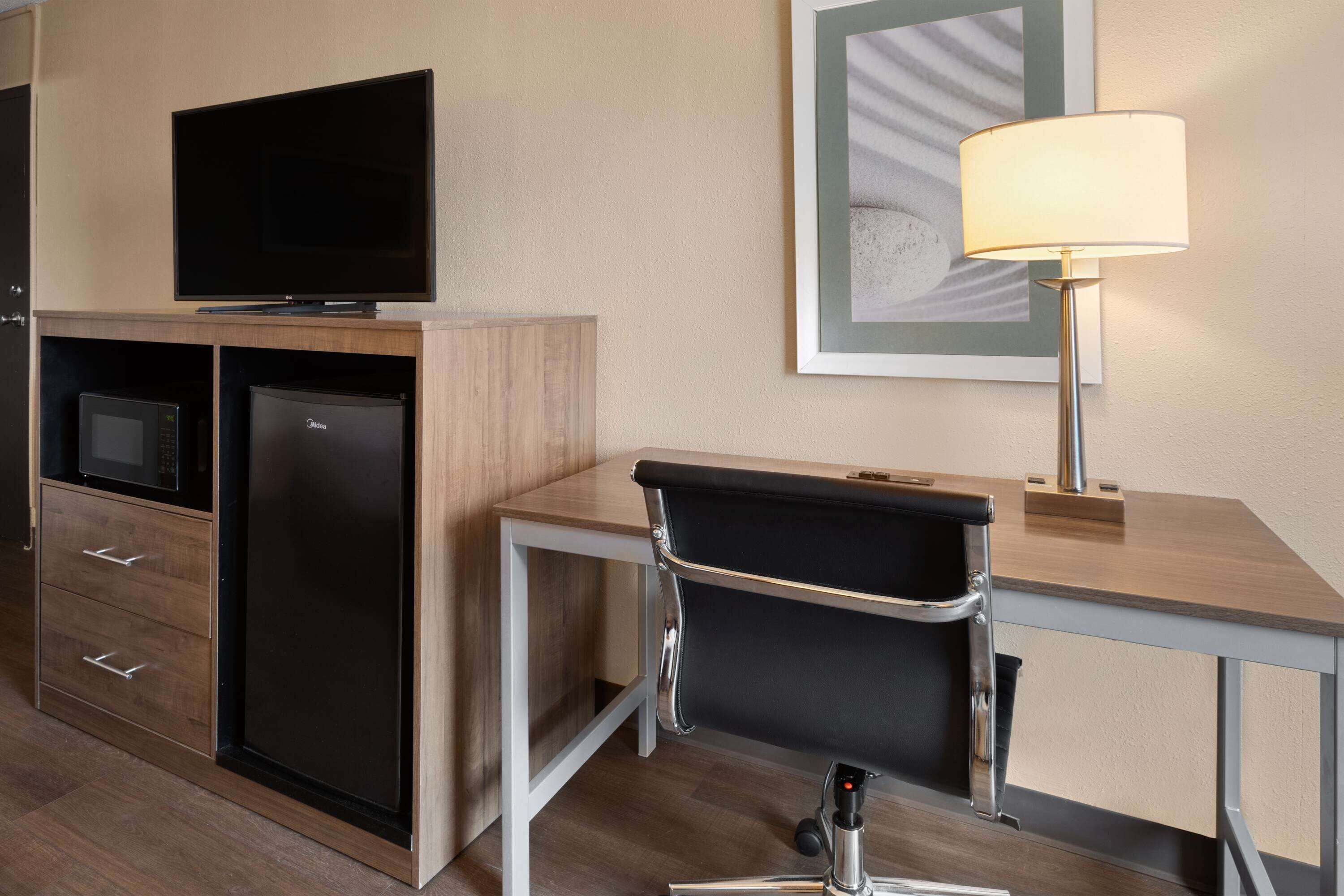 Ein kompaktes Hotelzimmer mit einer Küchenzeile, einem Flachbildfernseher, einem Schreibtisch mit Lampe und einem gerahmten Kunstwerk an der Wand.