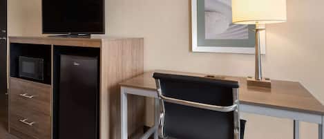 A compact hotel room with a kitchenette, a flat-screen TV, a desk with a lamp, and a framed artwork on the wall.