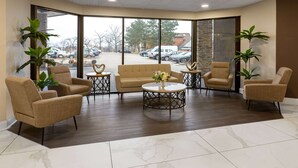Eine Lobby mit einem beigefarbenen Sofa, Sesseln und einem Couchtisch. Große Fenster bieten Blick auf parkende Autos und Gebäude.
