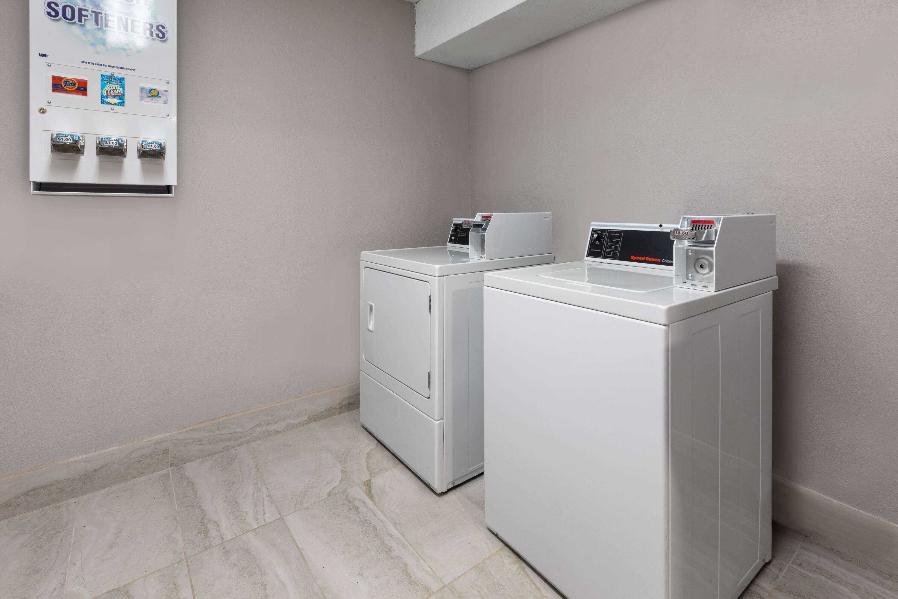 A laundry area with two white washers and dryers, a wall-mounted softener dispenser, and a tiled floor.