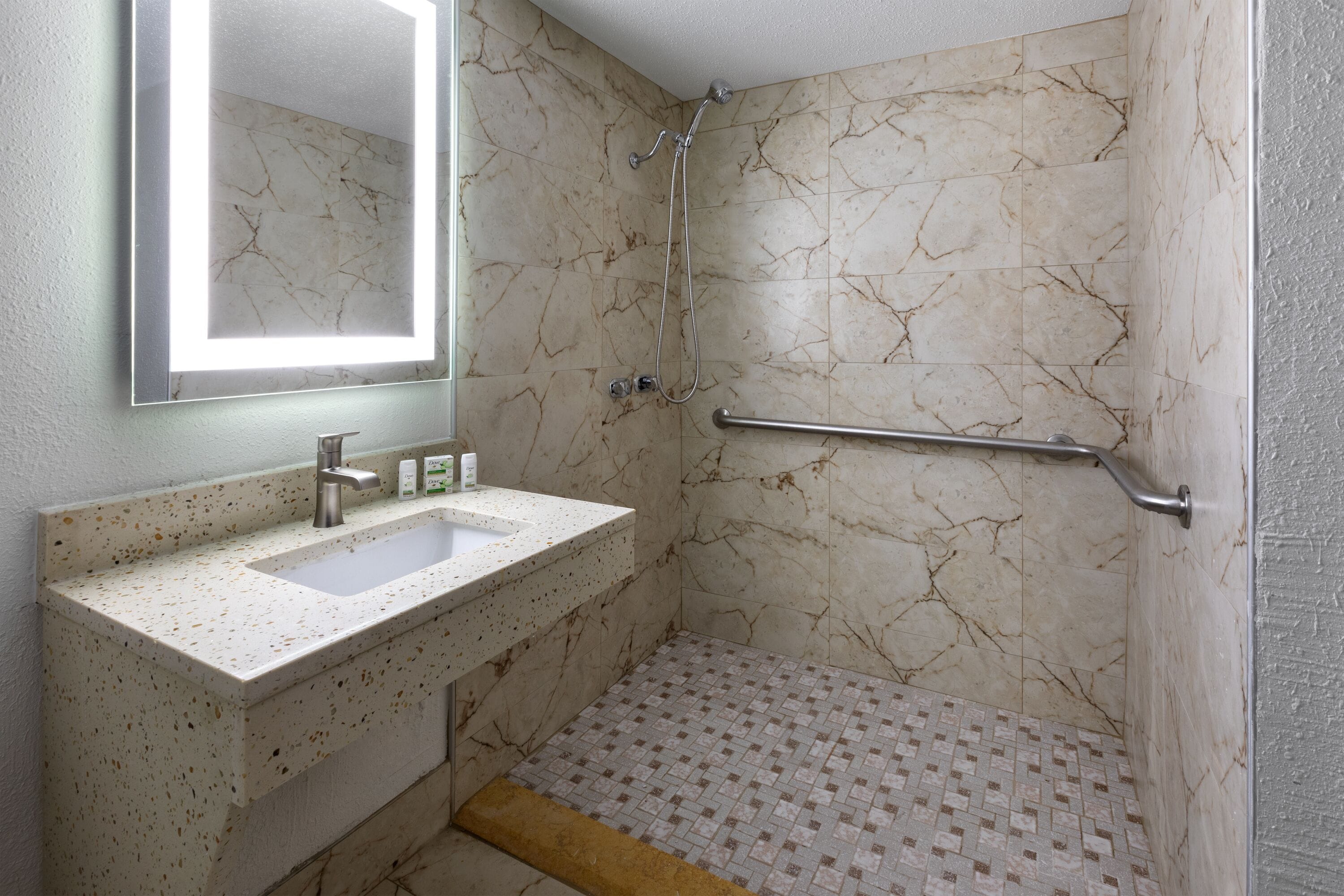 Une salle de bain avec une douche carrelée de marbre, un lavabo avec un miroir et une barre d’appui.