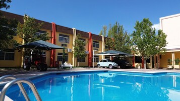 Una piscina con escalera metálica, rodeada por un edificio amarillo con detalles rojos, un coche blanco y un coche antiguo estacionado cerca.