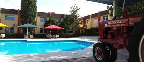 Piscina com um trator vermelho estacionado perto, rodeada por edifícios com fachadas amarelas e vermelhas.