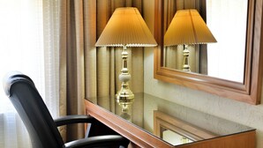 A hotel room with a wooden desk, two lamps, a chair, and a mirror reflecting the room.