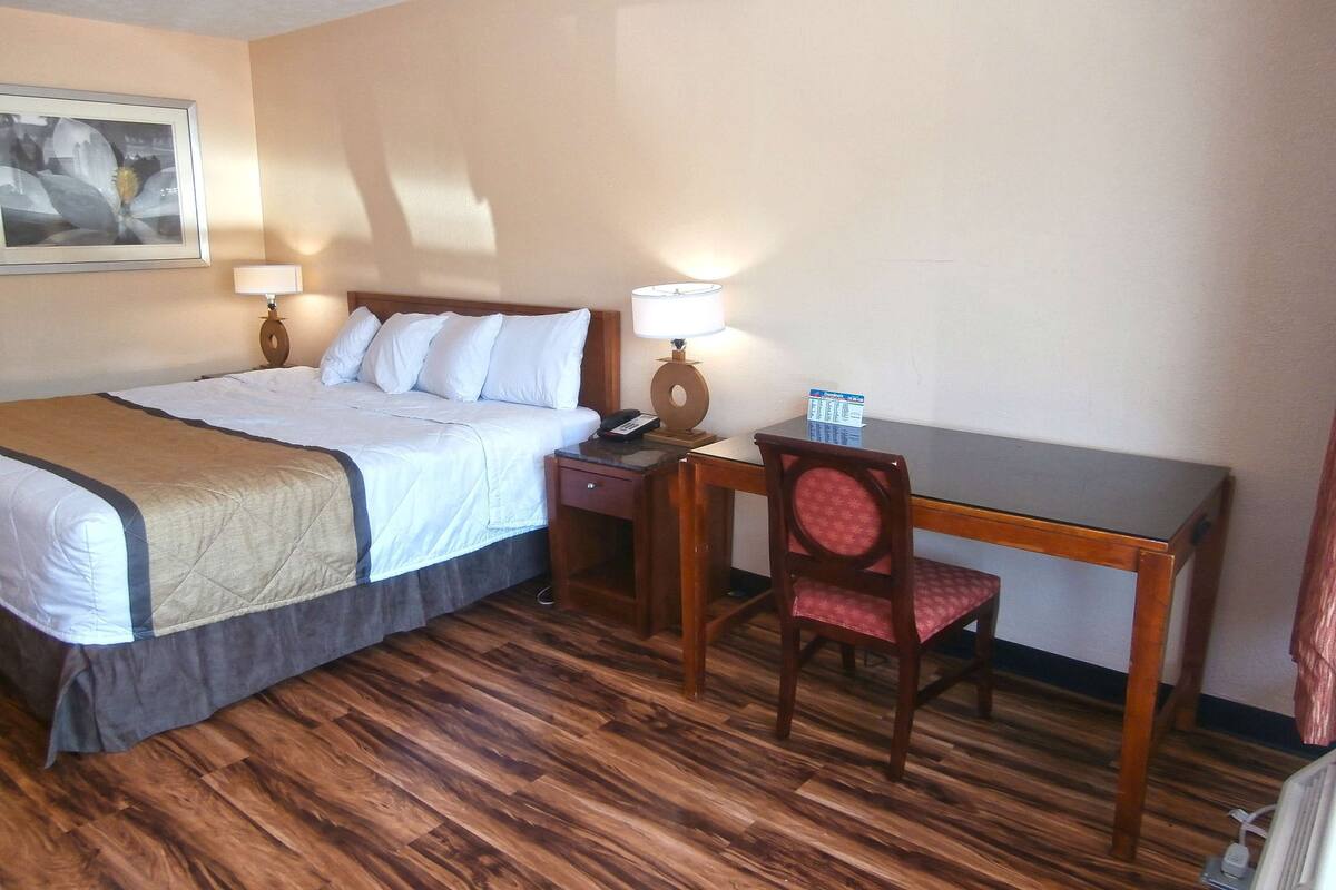 Ein Hotelzimmer mit Bett, Nachttisch, Lampe, Schreibtisch, Stuhl und einem Gemälde an der Wand.