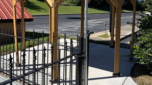 Um gazebo com telhado de metal e vigas de madeira, um portão de metal e um estacionamento ao fundo.