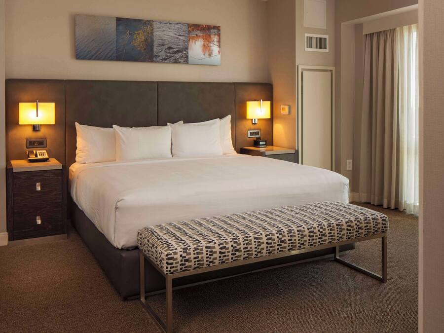 Une chambre d’hôtel avec un grand lit, deux tables de chevet avec des lampes, un banc au pied du lit et un tableau au mur.
