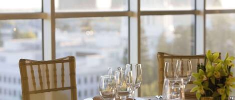 Una mesa de comedor dispuesta con platos, cubiertos y copas de vino, con vistas a edificios a través de amplios ventanales.