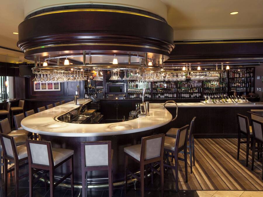 Uma área de bar com balcão curvo, vários banquinhos e uma variedade de garrafas e copos nas prateleiras.