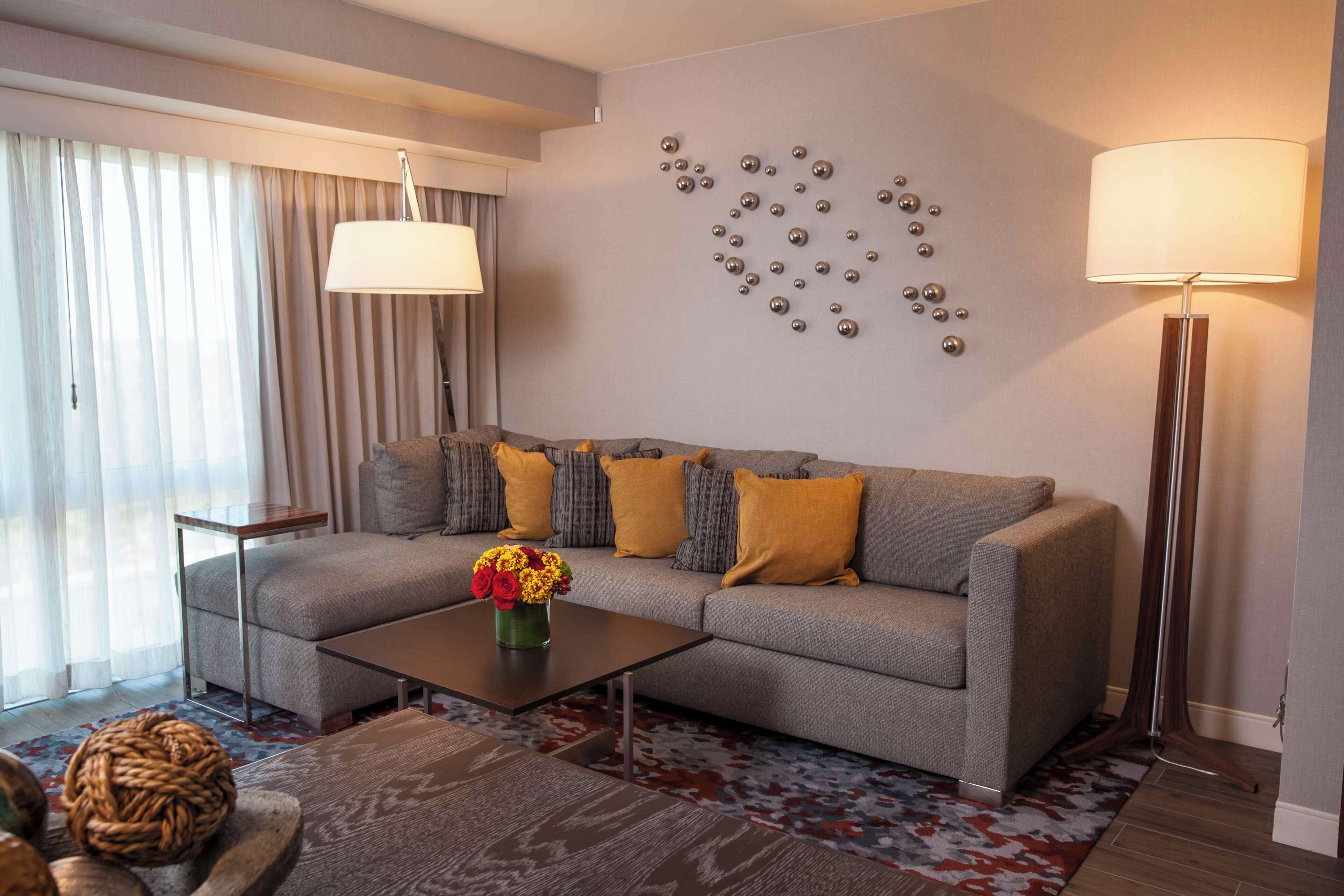 Una sala moderna con un sofá gris, cojines amarillos, una mesa de centro con un jarrón de flores, una alfombra estampada y una obra de arte en la pared.