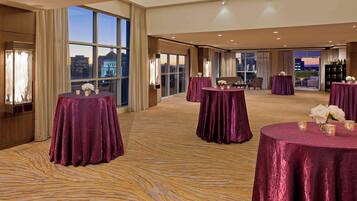 A spacious hotel room with large windows offering a city view, multiple round tables with purple tablecloths, and a carpeted floor.