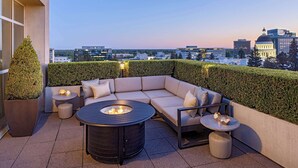 Una terraza en la azotea con un sofá seccional color beige, una mesa redonda con una chimenea y una mesita redonda con adornos.