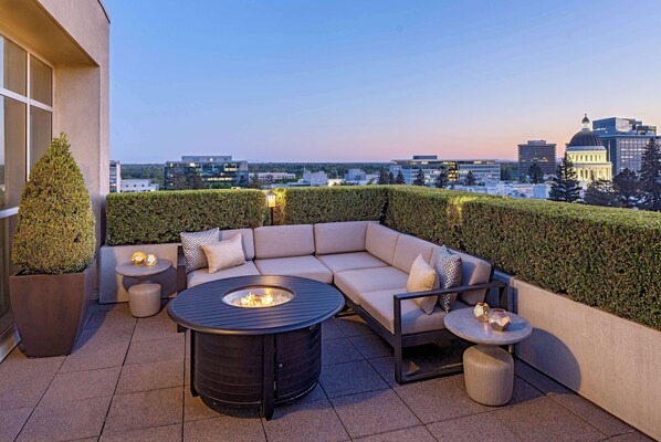A rooftop patio with a beige sectional sofa, a round table with a fire pit, and a small round table with decorative items.